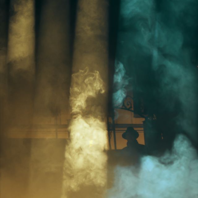 En rökridå som är upplyst i gult och grönt där man kan skymta en siluett av en man i bakgrunden.